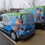 Two EnviroClean Industries vans wrapped in colorful graphics featuring a cartoon pressure washer character, company name, services, and contact details